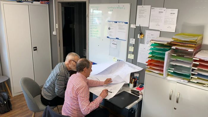 Dwóch mężczyzn analizuje rozłożony rysunek techniczny na biurku w biurze, na ścianie tablica z harmonogramem i segregatory