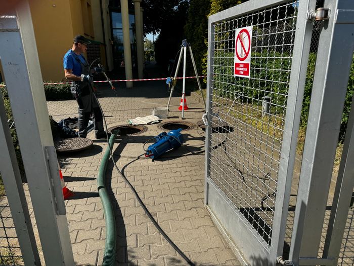 Pracownik z wężem przy otwartych włazach na kostce brukowej, obok pompa zanurzeniowa i trójnóg, teren wygrodzony taśmą ostrzegawczą