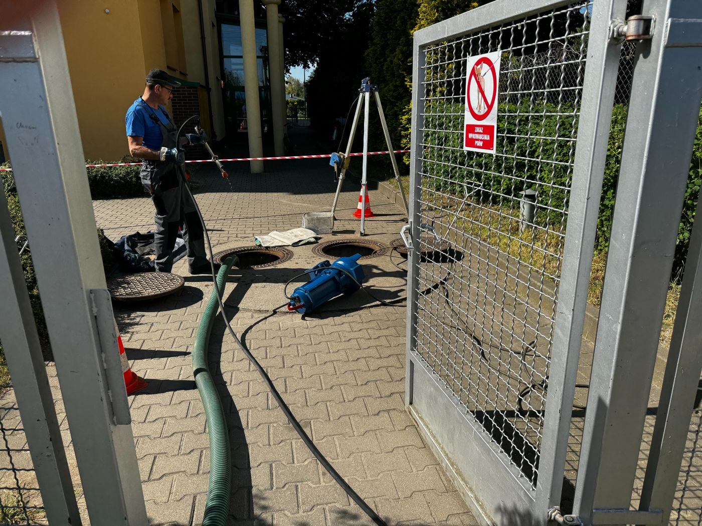 Pracownik z wężem przy otwartych włazach na kostce brukowej, obok pompa zanurzeniowa i trójnóg, teren wygrodzony taśmą ostrzegawczą