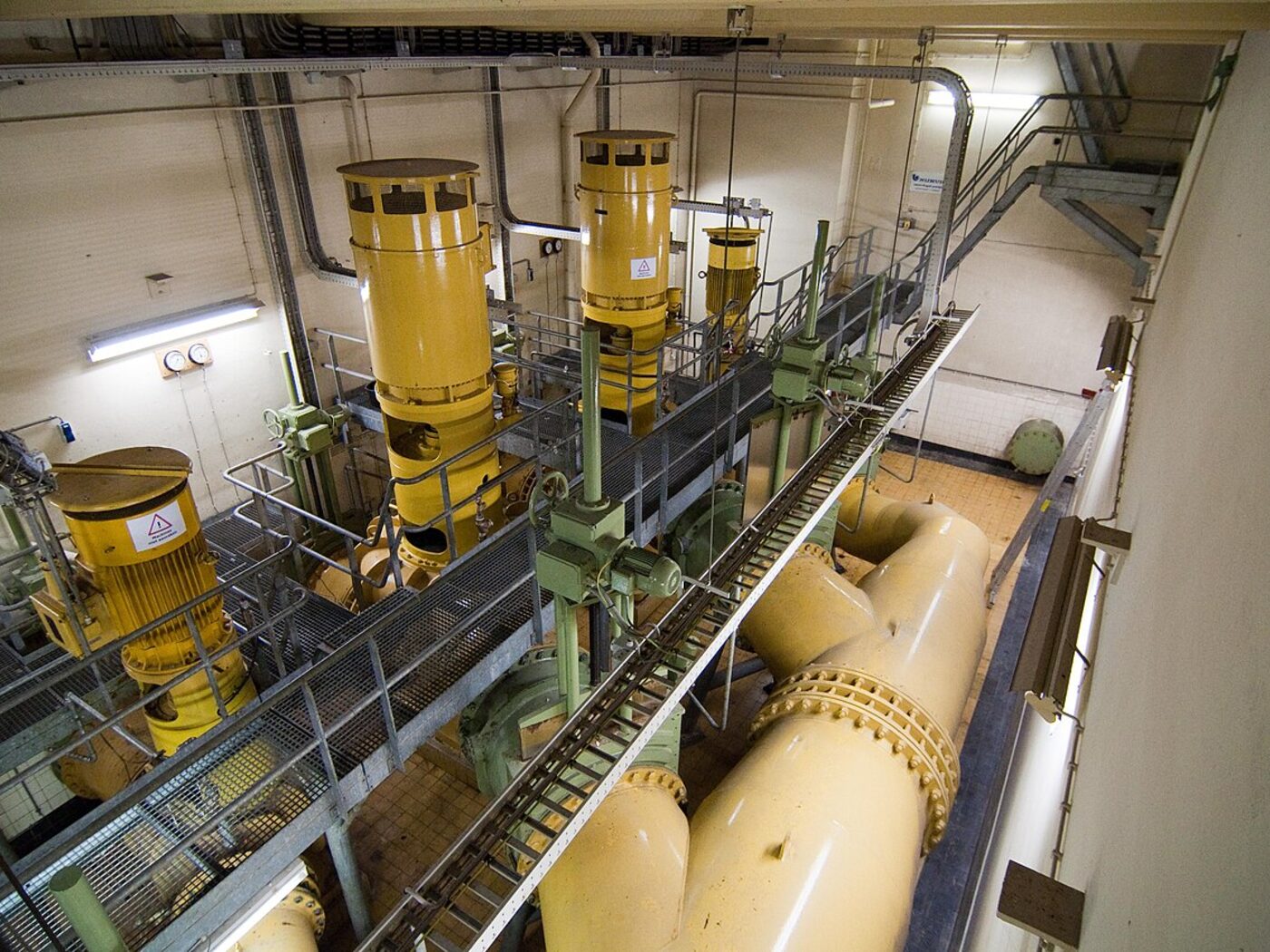 Hala technologiczna oczyszczalni ścieków: rząd żółtych zbiorników procesowych, rurociągi i pomosty technologiczne