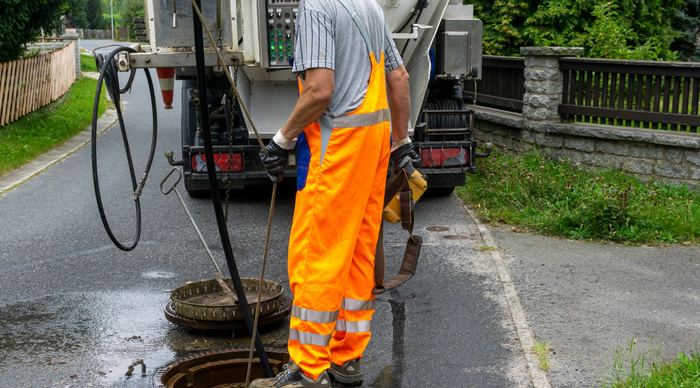 Pracownik w pomarańczowym ubraniu roboczym przy otwartej studzience z tyłu wozu ciśnieniowo-ssącego, w dłoni wąż wysokociśnieniowy