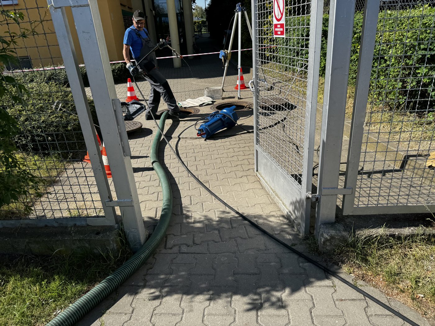 Pracownik obsługujący pompę zanurzeniową przy otwartych włazach przepompowni na terenie ogrodzonego obiektu, węże ssawne prowadzone przez furtkę